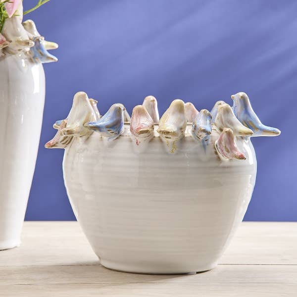 Ein Blumentopf von Werner Voß in Weiß mit bunten Vögeln (Ø 21, H 15,5 cm) steht auf einer hellen Holzfläche vor einem lilafarbenen Hintergrund. Dieser stilvolle Blumentopf ist ein dekoratives Highlight für jedes Zuhause.