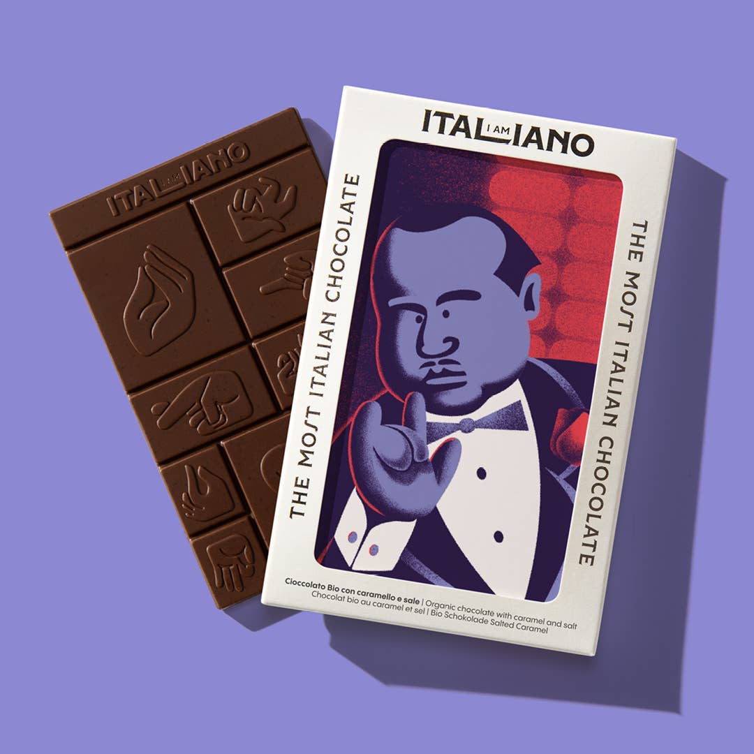 Eine 'Padrino' Bio-Schokolade mit Karamell und Salz von I AM ITALIANO, verziert mit italienischen Handgesten, liegt neben der Box. Auf der Verpackung stehen "I AM ITALIANO" und "The Most Italian Chocolate".