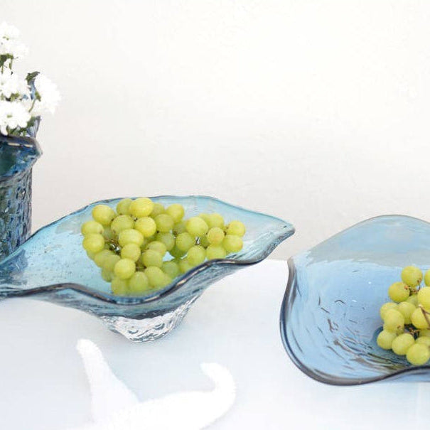 Zwei dekorative blaue Glasschalen von AULICA (33 x 18 x 10,5 cm), gefüllt mit grünen Weintrauben, stehen auf einer weißen Fläche. Links hält eine blaue Vase weiße Blumen, die teilweise im Rahmen sichtbar sind.
