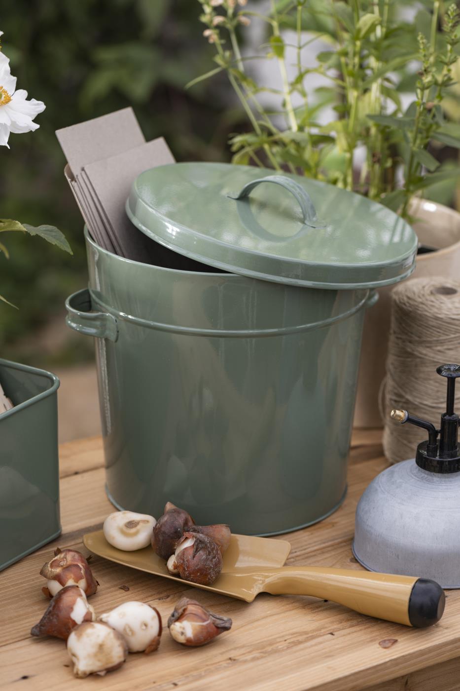 Ein grüner Metallbehälter mit Deckel steht auf einem Holztisch neben Samentütchen, der IB Laursen Kompakte Pflanzschaufel 'Clay', Blumenzwiebeln, Bindfaden, Sprühflasche und Topfpflanzen - ideales Gartenzubehör für jeden Gartenliebhaber.