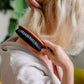 Eine Person mit blondem Haar in einem hellen Gewand bürstet ihr Haar mit der Croll & Denecke Haarbürste aus Birnenholz mit Naturborsten, wobei sie ihr Haar hochhält und die plastikfreie Bürste verwendet, die von hinten und von der Seite sichtbar ist.