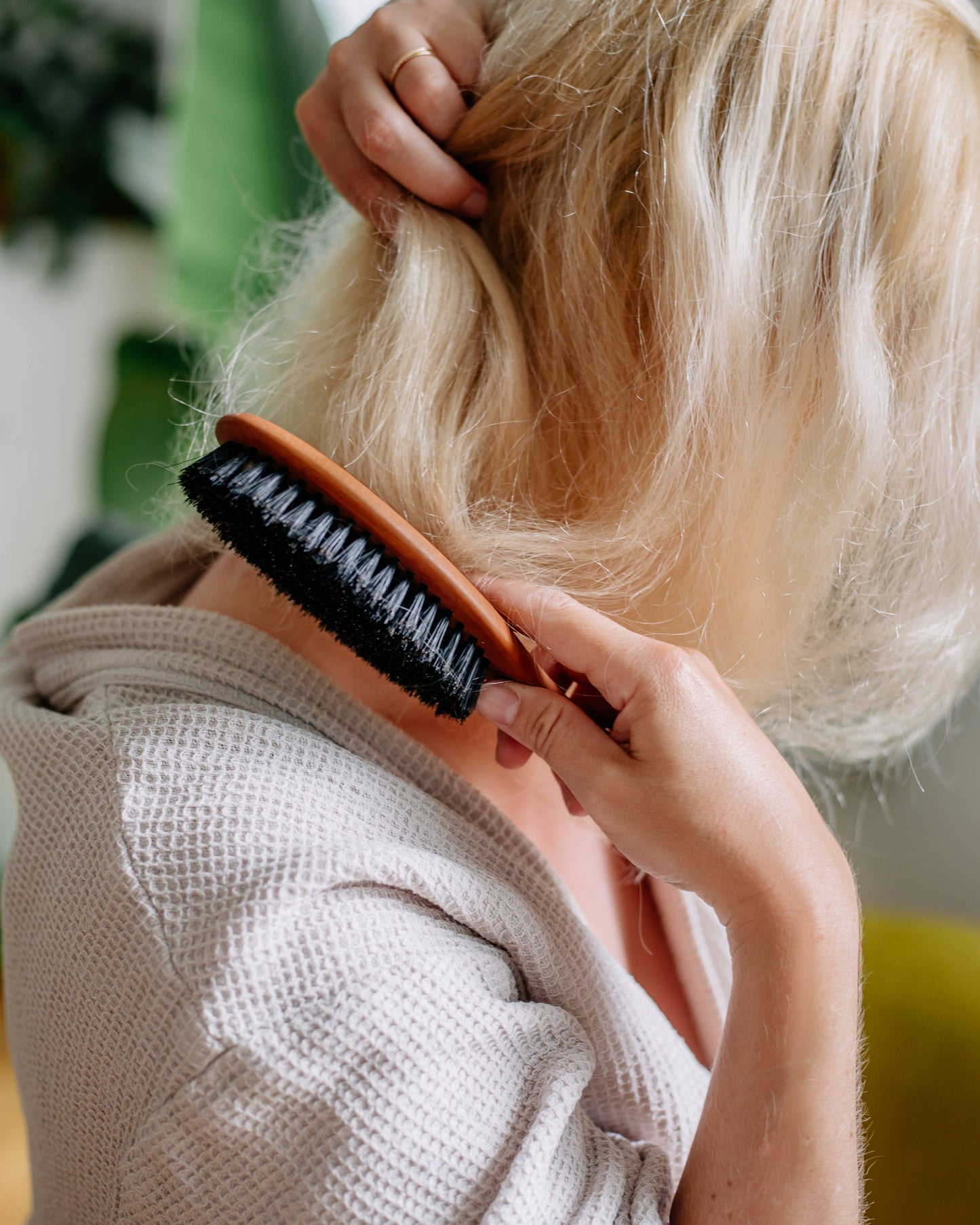 Eine Person mit blondem Haar in einem hellen Gewand bürstet ihr Haar mit der Croll & Denecke Haarbürste aus Birnenholz mit Naturborsten, wobei sie ihr Haar hochhält und die plastikfreie Bürste verwendet, die von hinten und von der Seite sichtbar ist.
