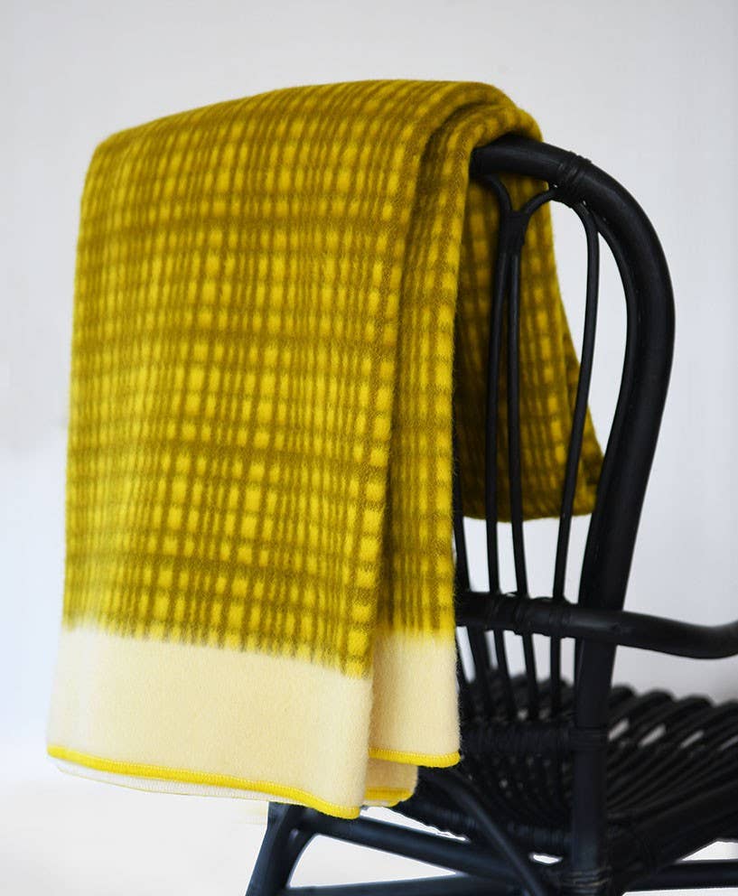 Eine Sofie Sjöström | 'Darling' Gelbe Tagesdecke aus Lammwolle von Sofie Sjöström Design ist über die Rückenlehne eines schwarzen Rattanstuhls vor einem schlichten weißen Hintergrund drapiert.