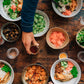 Draufsicht auf mehrere Schüsseln mit Reis, Gemüse und Beilagen – einige davon garniert mit „White Mausu | Black Bean Rayu Sauce“ von White Mausu Ltd und einem Spritzer scharfer Soße – auf einem rustikalen Tisch, wobei Hände nach dem Essen und den Soßen greifen. Außerdem stehen Gläser mit Wasser, kleine Beilagen und Schüsseln mit herzhaften fermentierten Bohnen bereit.