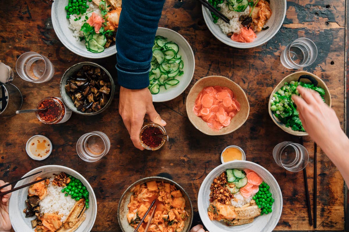 Draufsicht auf mehrere Schüsseln mit Reis, Gemüse und Beilagen – einige davon garniert mit „White Mausu | Black Bean Rayu Sauce“ von White Mausu Ltd und einem Spritzer scharfer Soße – auf einem rustikalen Tisch, wobei Hände nach dem Essen und den Soßen greifen. Außerdem stehen Gläser mit Wasser, kleine Beilagen und Schüsseln mit herzhaften fermentierten Bohnen bereit.