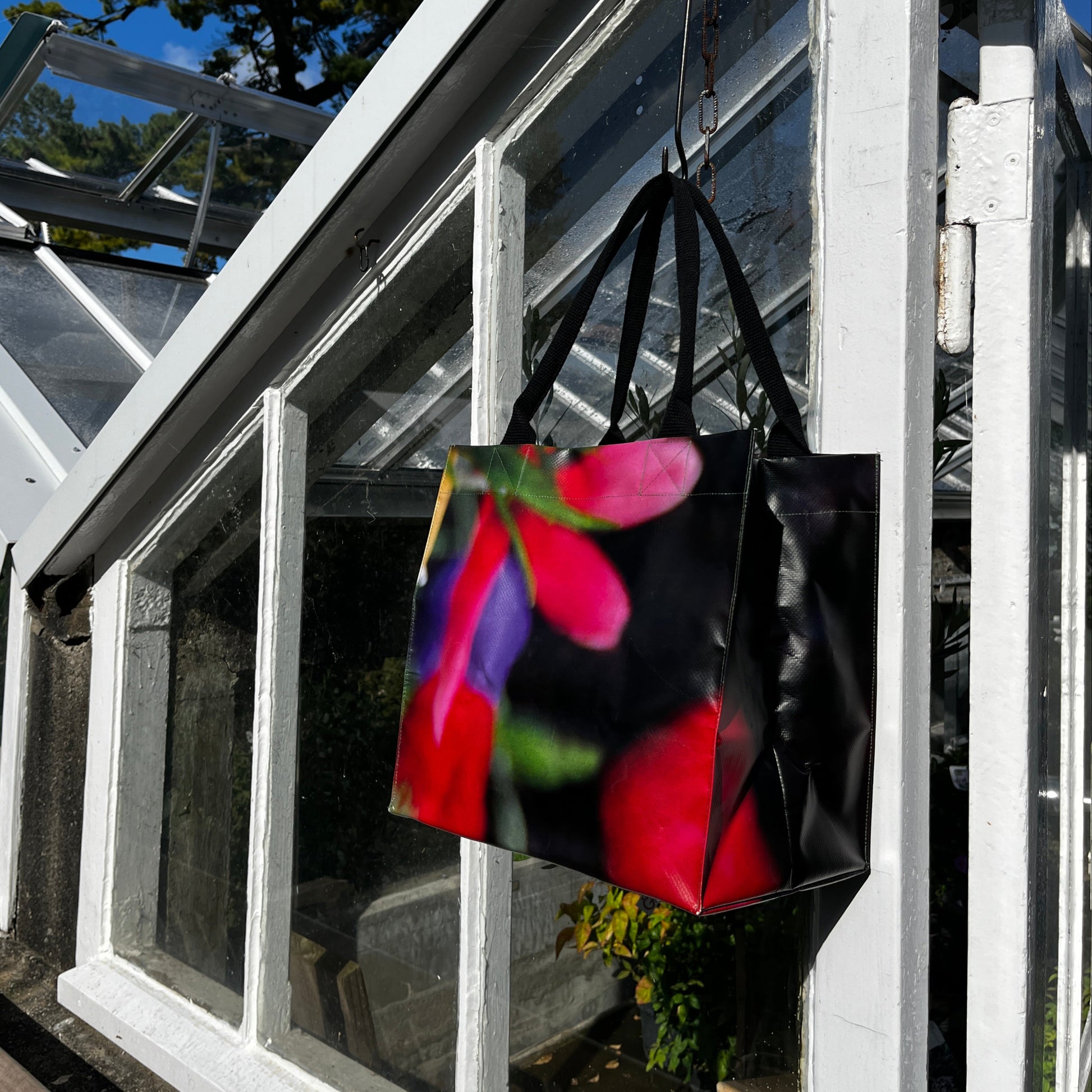 Die KGA Upcycling Tasche 'Blume' der Königlichen Gartenakademie mit ihrem lebendigen Blumenmuster hängt an einer weiß gerahmten Gewächshaustür vor einem Hintergrund aus blauem Himmel und grünen Bäumen.