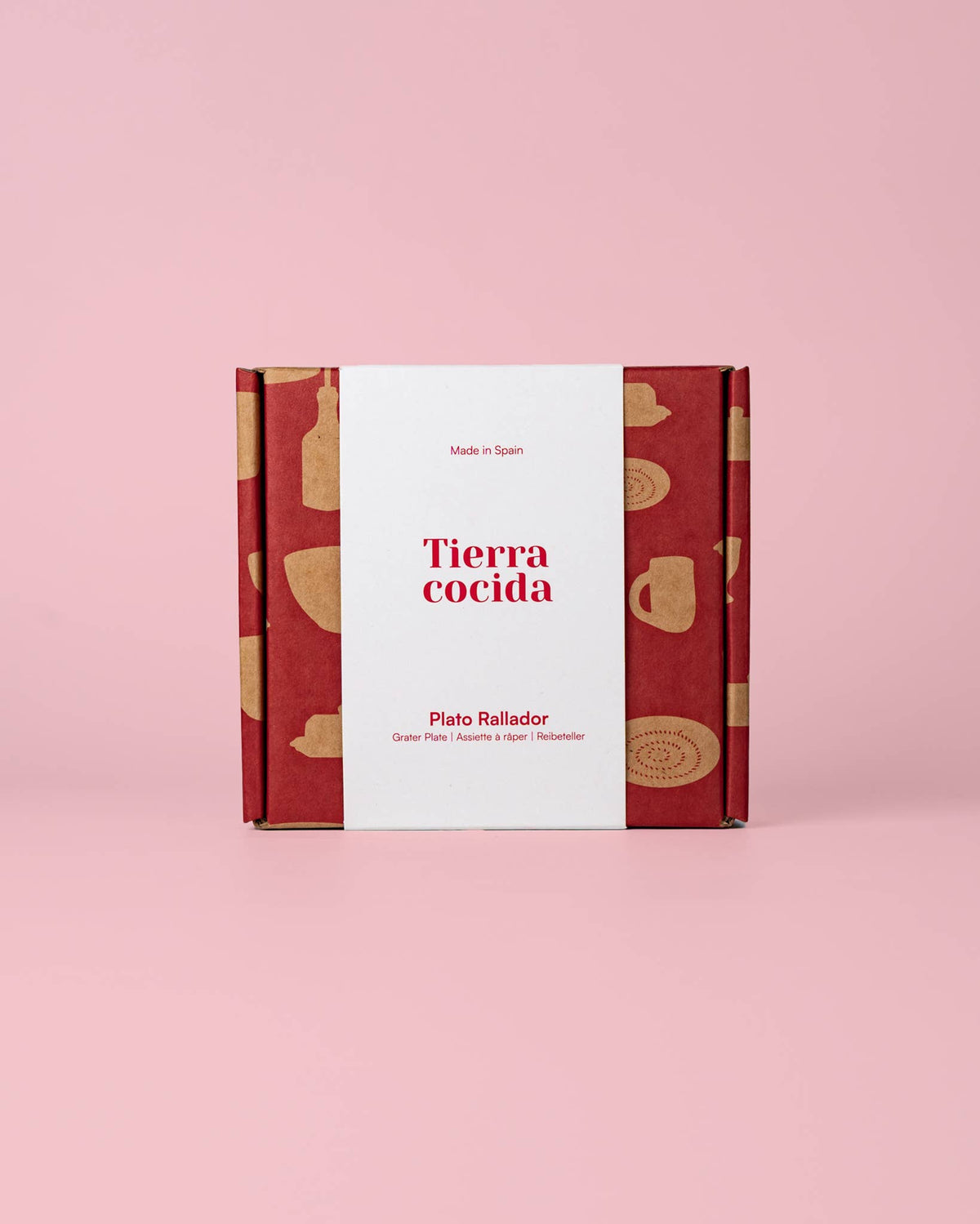 Auf einem rosa Hintergrund steht eine aufrecht stehende rot-beige Schachtel mit einem weißen Etikett, auf dem „Alfar Tierra Cocida | Kleine Keramik Knoblauchreibe ‚Pomodoro‘“ zu lesen ist. Die Verpackung zeigt Illustrationen von beigen Küchenutensilien und unterstreicht damit die traditionelle Keramikkunst dieser handgefertigten spanischen Knoblauchreibe von Alfar Tierra Cocida.