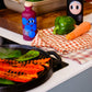 Bunte Flaschen, eine grüne Paprika und eine Karotte liegen auf einem karierten Tuch neben einer Grillpfanne mit rotem Paprika und Grünzeug. Im Hintergrund bereitet jemand Blattgemüse neben einer Flasche Yiayia and Friends | Gereifter Balsamico-Essig 200 ml zu.
