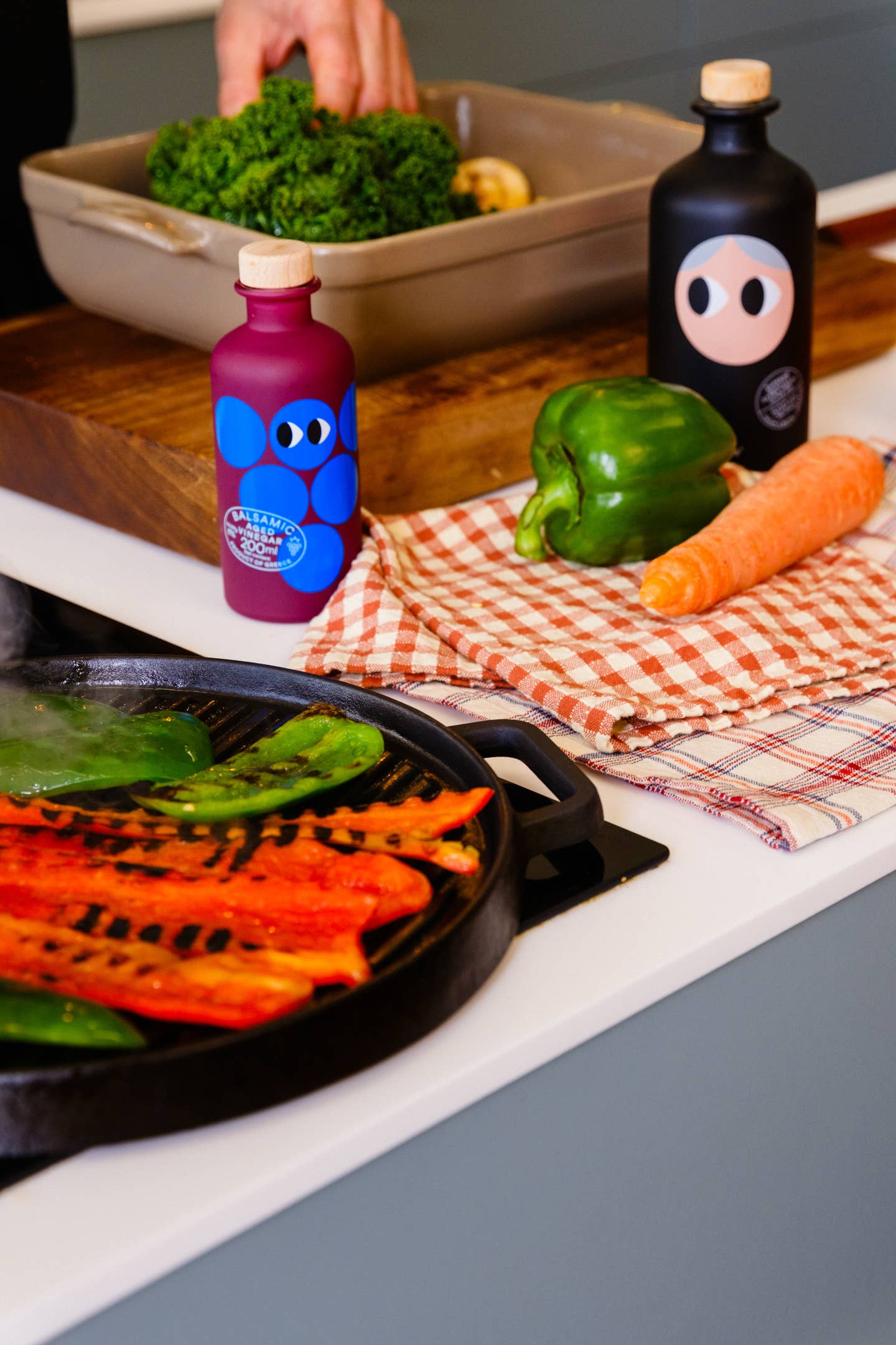 Bunte Flaschen, eine grüne Paprika und eine Karotte liegen auf einem karierten Tuch neben einer Grillpfanne mit rotem Paprika und Grünzeug. Im Hintergrund bereitet jemand Blattgemüse neben einer Flasche Yiayia and Friends | Gereifter Balsamico-Essig 200 ml zu.