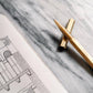 Der ENTREPÔT YSTUDIO Klassischer Revolve-Rollerball-Stift (Messing) liegt auf grauem Marmor neben einem Skizzenbuch mit architektonischer Zeichnung und bietet sanftes Schreiben sowie einen stabilen Griff für präzise Kreativität.