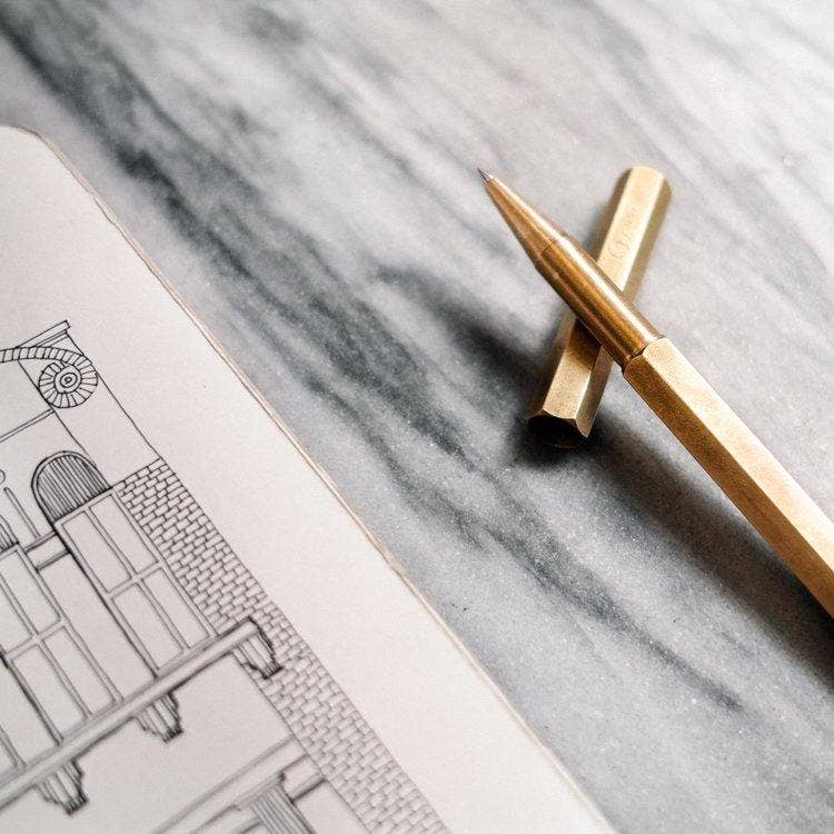 Der ENTREPÔT YSTUDIO Klassischer Revolve-Rollerball-Stift (Messing) liegt auf grauem Marmor neben einem Skizzenbuch mit architektonischer Zeichnung und bietet sanftes Schreiben sowie einen stabilen Griff für präzise Kreativität.