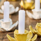 Weiße Kerzen leuchten in gelben und weißen blumenförmigen Keramikhaltern, die mit dem Ib Laursen Kerzenhalter aus Keramik Narzisse 'Buttercreme', von IB Laursen, auf Holz arrangiert, für einen frischen Frühlingsdeko Steinzeug Look sorgen.
