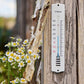 Ein IB Laursen Thermometer aus Emaille von IB Laursen ist an einem Holzpfosten in der Nähe eines Zweigs und kleiner weißer Gänseblümchen angebracht und zeigt ungefähr 18°C an. Der Hintergrund erscheint unscharf.