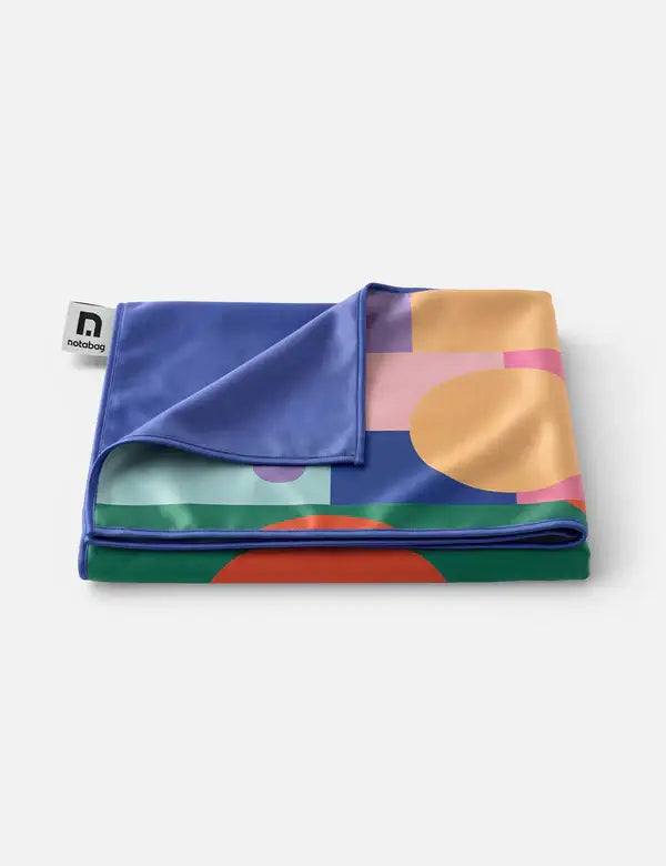 Ein gefaltetes Notabag 'Flow' Handtuch (78 x 160 cm) von Notabag mit einem leuchtenden geometrischen Druck in Blau, Orange, Grün und Rosa und einer einfarbig blauen Unterseite. Hergestellt aus recyceltem Handtuch-Material und mit einem Notabag-Anhänger an einer Ecke versehen.