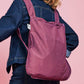 Eine Person in einer Jeansjacke trägt die Notabag | Wandelbare Tasche und Rucksack „Pflaume“ von Notabag auf dem Rücken vor einem rosa Hintergrund.