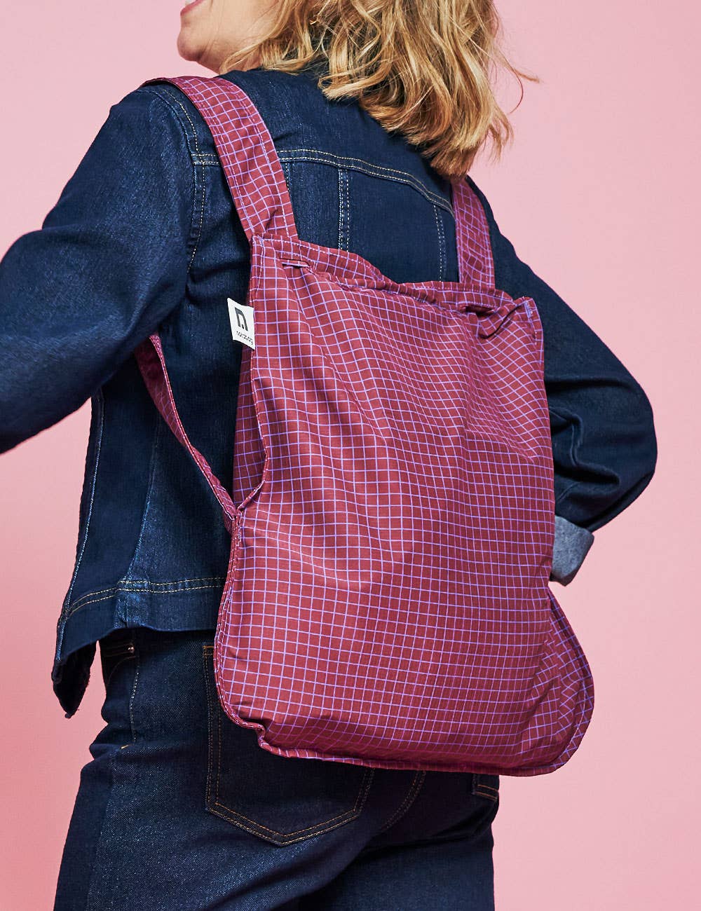 Eine Person in einer Jeansjacke trägt die Notabag | Wandelbare Tasche und Rucksack „Pflaume“ von Notabag auf dem Rücken vor einem rosa Hintergrund.