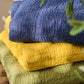 Drei gefaltete, strukturierte Quilts in Blau, Gelb und Hellgrün - darunter die IB Laursen Quilt Decke hellgrün mit Kreuzstich - sind auf Holz gestapelt, davor eine Blattpflanze, ideal für skandinavische Wohndeko.