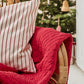 Ein Korbsessel mit einer IB Laursen Quilt Decke rot mit Kreuzstich und einem weißen Kissen mit roten Streifen steht vor einem mit Ornamenten und Lichtern geschmückten Weihnachtsbaum.