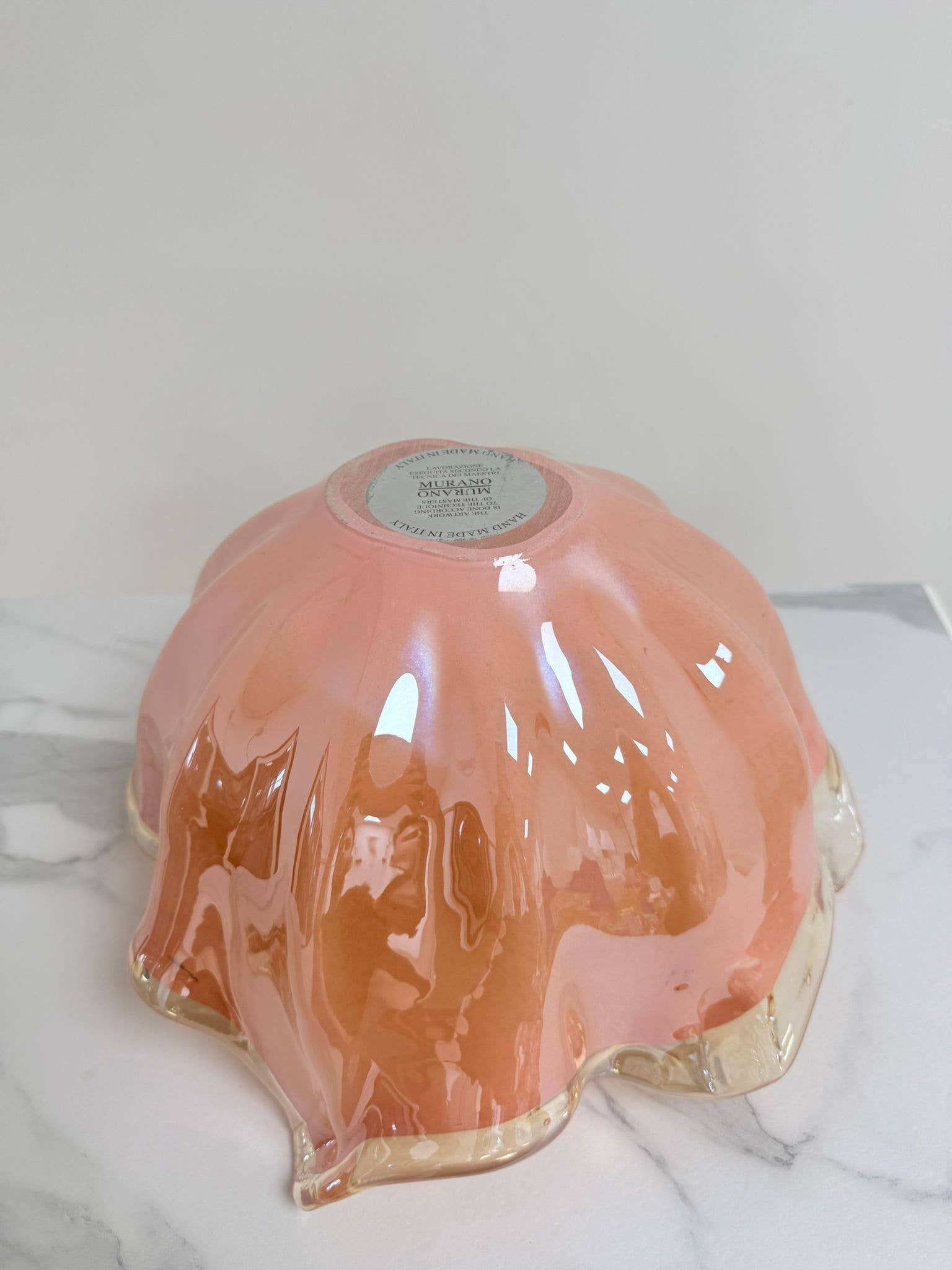 Die Murano Dream Dekoschale „Fazzoletto“ aus rosa Murano-Glas (H 20, Ø 22 cm) steht kopfüber auf weißem Marmor und präsentiert dabei ihre gewellten Ränder und das Etikett am Boden, während ihre glänzende Oberfläche das Licht und die Umgebung reflektiert.