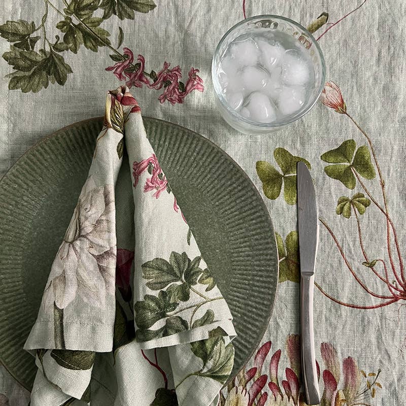 Ein grüner, gerippter Teller, eine Leinenserviette von Koustrupco mit dem Motiv „Flora Danica Atlas“ (45 × 45 cm), ein Glas Eiswasser und ein Messer liegen auf einem Tisch mit einer Tischdecke mit botanischem Muster.
