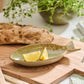 Die Ib Laursen Schale 'Ocean Green' in Fischform steht mit zwei Zitronenspalten auf einem Holzbrett; dahinter liegen ein Brotlaib, frische Kräuter, Küchenutensilien und ein gestreiftes Tuch.
