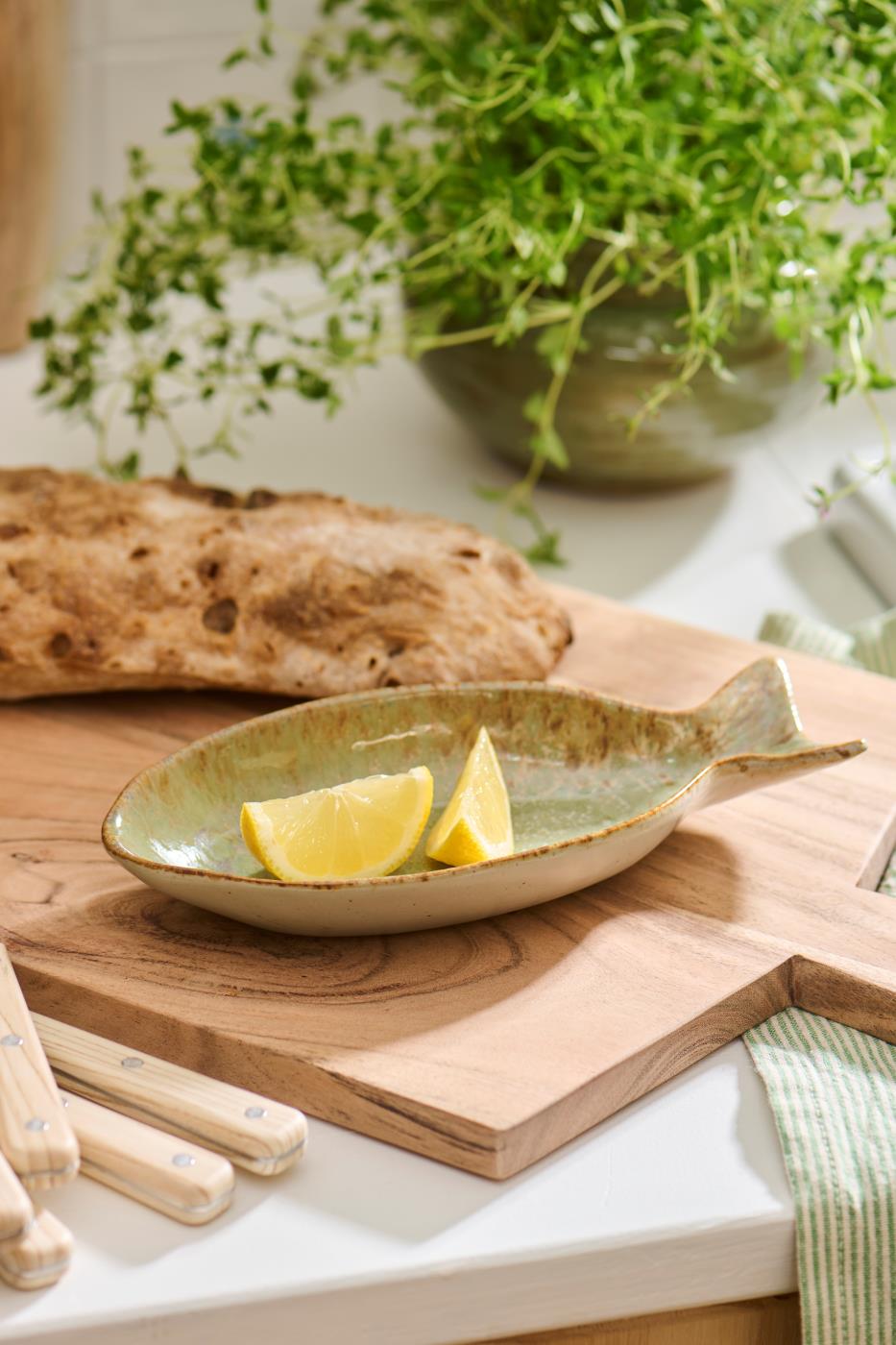 Die Ib Laursen Schale 'Ocean Green' in Fischform steht mit zwei Zitronenspalten auf einem Holzbrett; dahinter liegen ein Brotlaib, frische Kräuter, Küchenutensilien und ein gestreiftes Tuch.