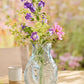Die Ib Laursen Vase fischförmig in Hellblau, 24 cm hoch von Königliche Gartenakademie, präsentiert lila, pinke und grüne Blumen auf einem Holztisch mit einer Tasse und verschwommenem Garten im Hintergrund.