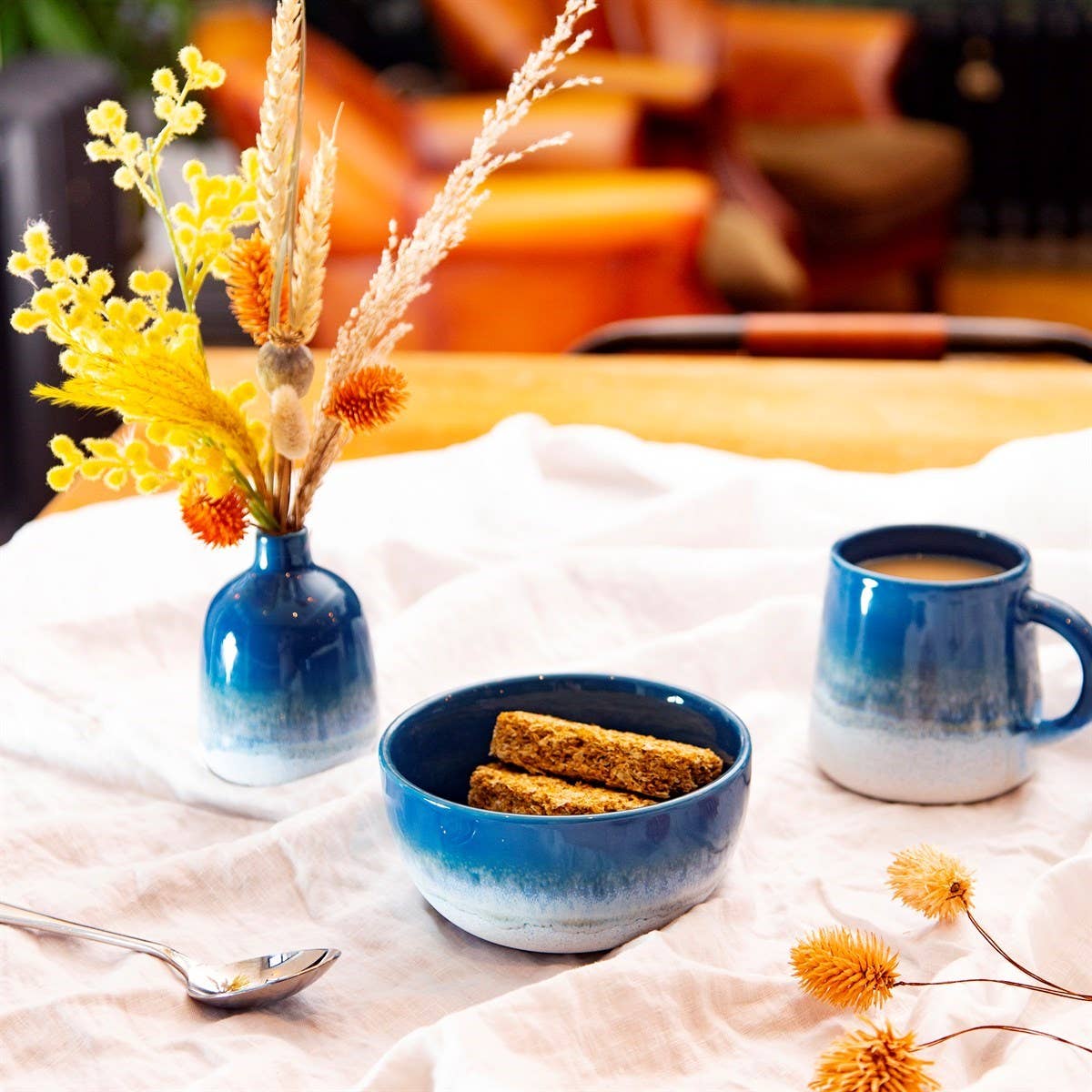 Eine blaue Schüssel mit Müsliriegeln, eine blaue Tasse in einem ozeanblauen Farbton, ein Löffel und die blaue Vase „Mojave Glaze“ von Sass & Belle Europe (12,5 cm), gefüllt mit gelben und orangefarbenen Blumen, sind auf einem weißen Tischtuch im Boho-Stil arrangiert, im Hintergrund sind verschwommene braune Stühle zu sehen.