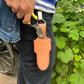 Eine Person in schwarzer Hose und gestreiftem Hemd hält eine Gartenschere neben einer Niwaki | 'Belt Clip' Praktische Halterung für Niwaki Single Holster von Niwaki, die an ihrem Gürtel befestigt ist, und steht in der Nähe von grünen Blattpflanzen.