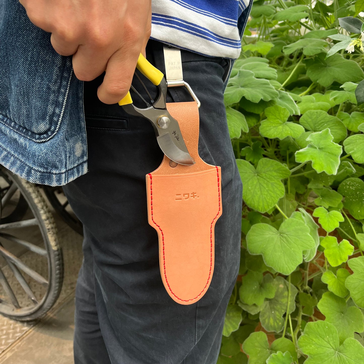 Eine Person in schwarzer Hose und gestreiftem Hemd hält eine Gartenschere neben einer Niwaki | 'Belt Clip' Praktische Halterung für Niwaki Single Holster von Niwaki, die an ihrem Gürtel befestigt ist, und steht in der Nähe von grünen Blattpflanzen.