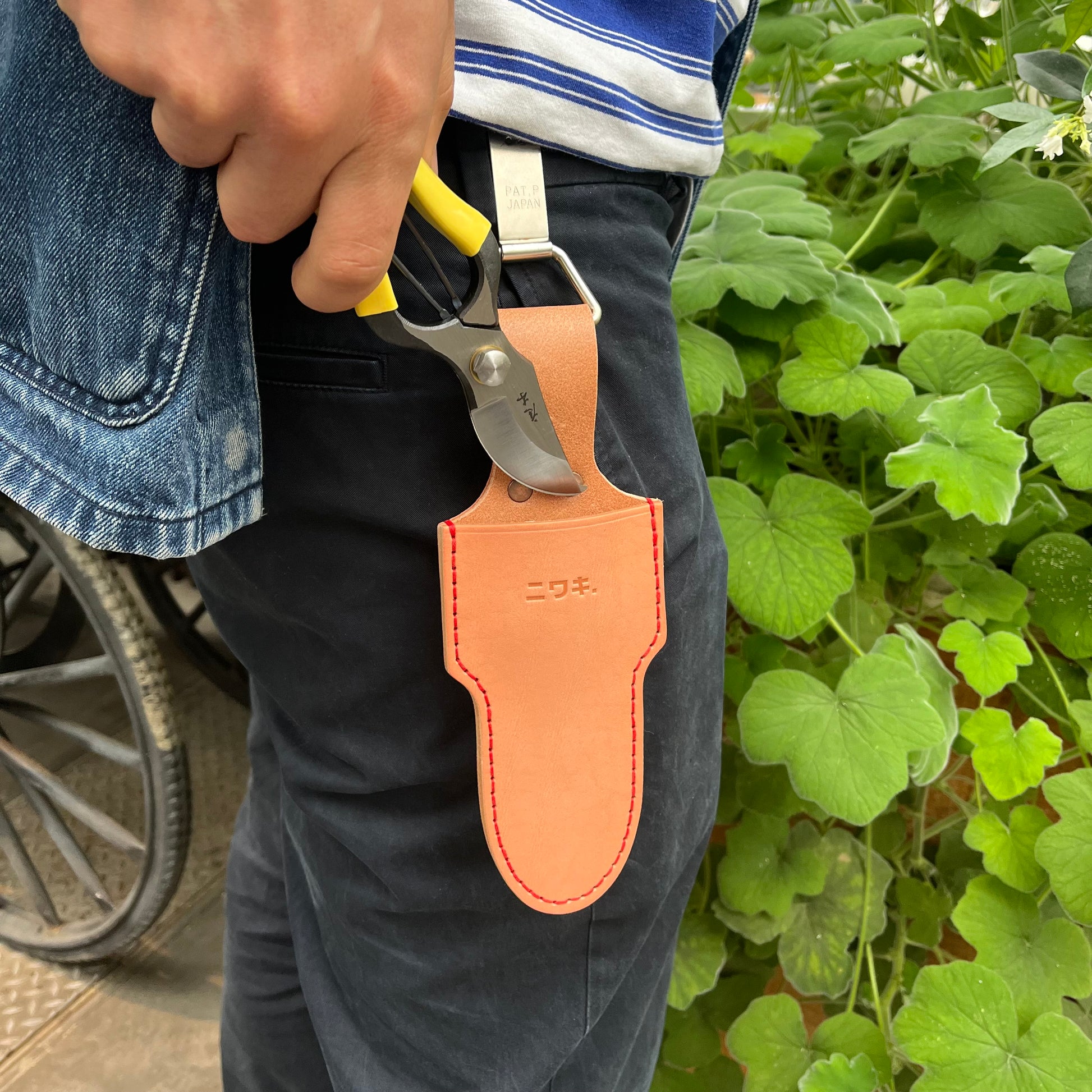 Eine Person in schwarzer Hose und gestreiftem Hemd hält eine Gartenschere neben einer Niwaki | 'Belt Clip' Praktische Halterung für Niwaki Single Holster von Niwaki, die an ihrem Gürtel befestigt ist, und steht in der Nähe von grünen Blattpflanzen.