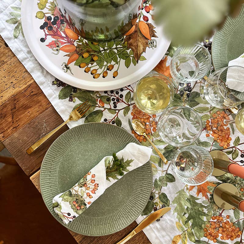 Ein gedeckter Tisch mit grünen Tellern, goldenem Besteck, Weingläsern sowie einer herbstlich gestalteten Tischdecke und Servietten mit Blumenmotiven, die mit Blättern und Beeren verziert sind, schafft eine festliche, gemütliche Atmosphäre. In der Mitte sorgt das Serviertablett „Herbst“ von Koustrupco (Ø 38 cm) für eine elegante Note – sein Design aus Birkenfurnier bringt das Herbstmotiv wunderschön zur Geltung und passt perfekt zur warmen saisonalen Dekoration.