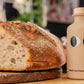 Ein rustikaler Brotlaib liegt auf einem Holzbrett neben einer beigen Flasche yiayia and friends Natives Olivenöl Extra mit Knoblauch 200 ml - ideal für die mediterrane Küche; im Hintergrund ist eine unscharfe schwarze Flasche zu sehen.