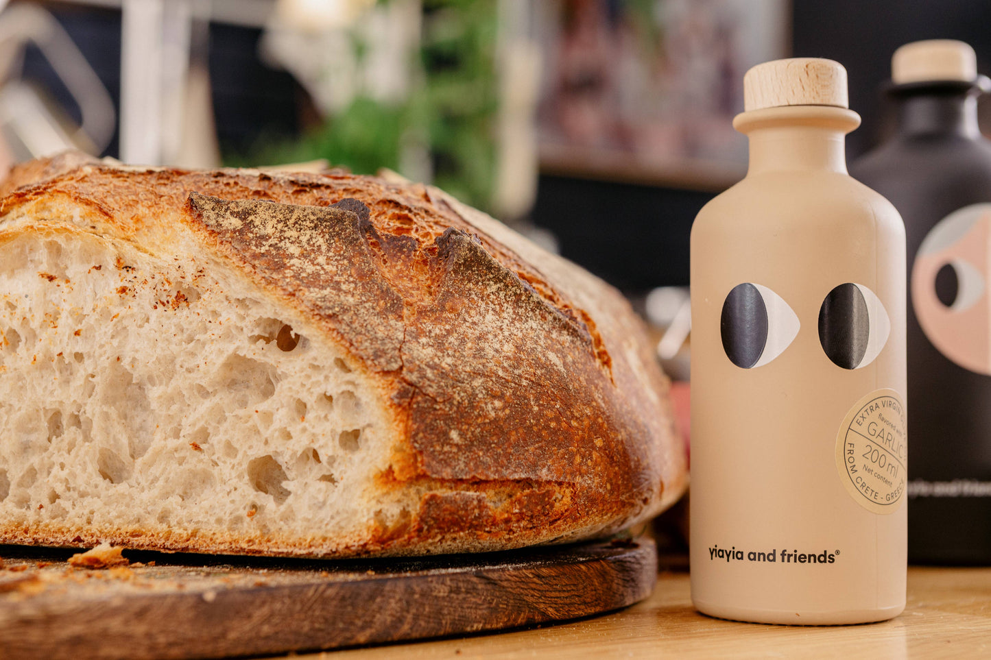 Ein rustikaler Brotlaib liegt auf einem Holzbrett neben einer beigen Flasche yiayia and friends Natives Olivenöl Extra mit Knoblauch 200 ml - ideal für die mediterrane Küche; im Hintergrund ist eine unscharfe schwarze Flasche zu sehen.