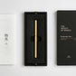 Ein goldener YSTUDIO | Klassischer Revolve-Rollerball-Stift (Messing) von ENTREPÔT mit stabilem Griff liegt in einer schwarzen Box, daneben ein weißes Booklet mit japanischen Zeichen und eine schwarze Hülle mit THE WEIGHT OF WORDS Classic Series.