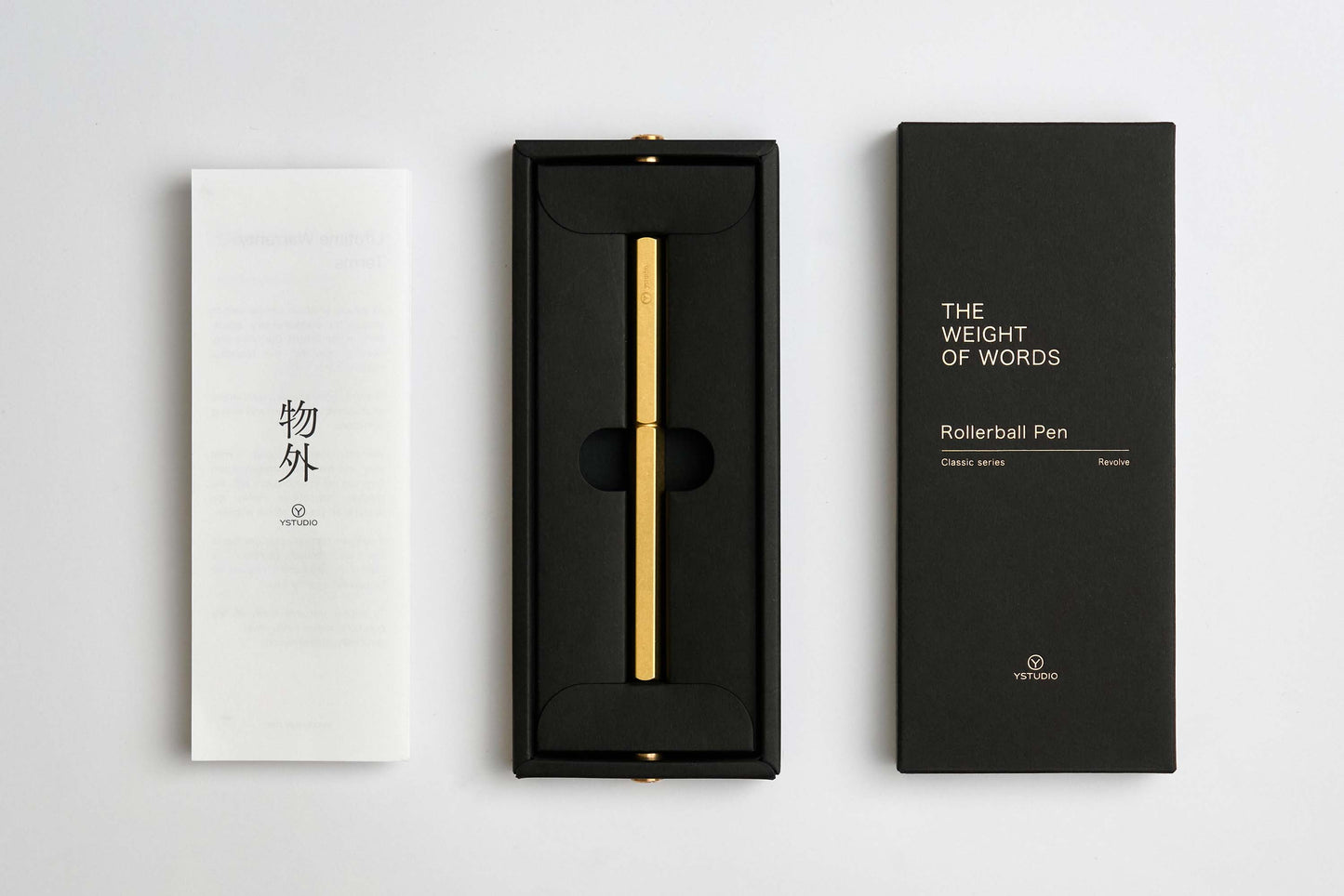 Ein goldener YSTUDIO | Klassischer Revolve-Rollerball-Stift (Messing) von ENTREPÔT mit stabilem Griff liegt in einer schwarzen Box, daneben ein weißes Booklet mit japanischen Zeichen und eine schwarze Hülle mit THE WEIGHT OF WORDS Classic Series.