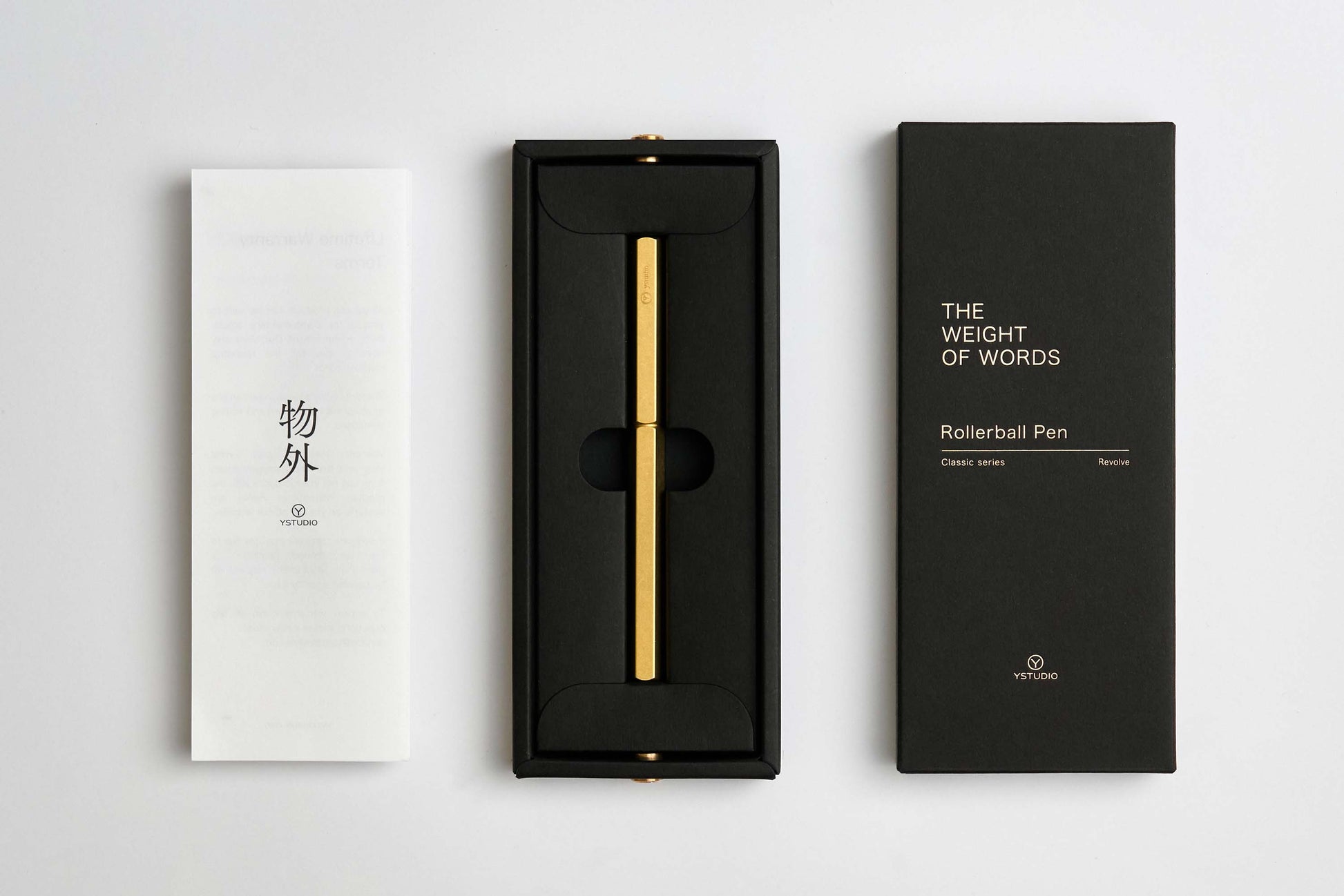 Ein goldener YSTUDIO | Klassischer Revolve-Rollerball-Stift (Messing) von ENTREPÔT mit stabilem Griff liegt in einer schwarzen Box, daneben ein weißes Booklet mit japanischen Zeichen und eine schwarze Hülle mit THE WEIGHT OF WORDS Classic Series.