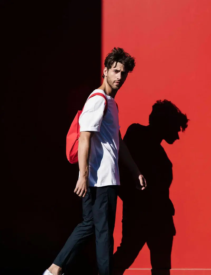 Ein Mann mit zerzaustem Haar, der ein weißes T-Shirt und eine dunkle Hose trägt, hält die Notabag | „Original Red“ – Tasche und Rucksack in einem von Notabag in der Hand, während er an einer roten Wand vorbeigeht und dabei einen deutlichen Schatten auf den hellen Hintergrund wirft.