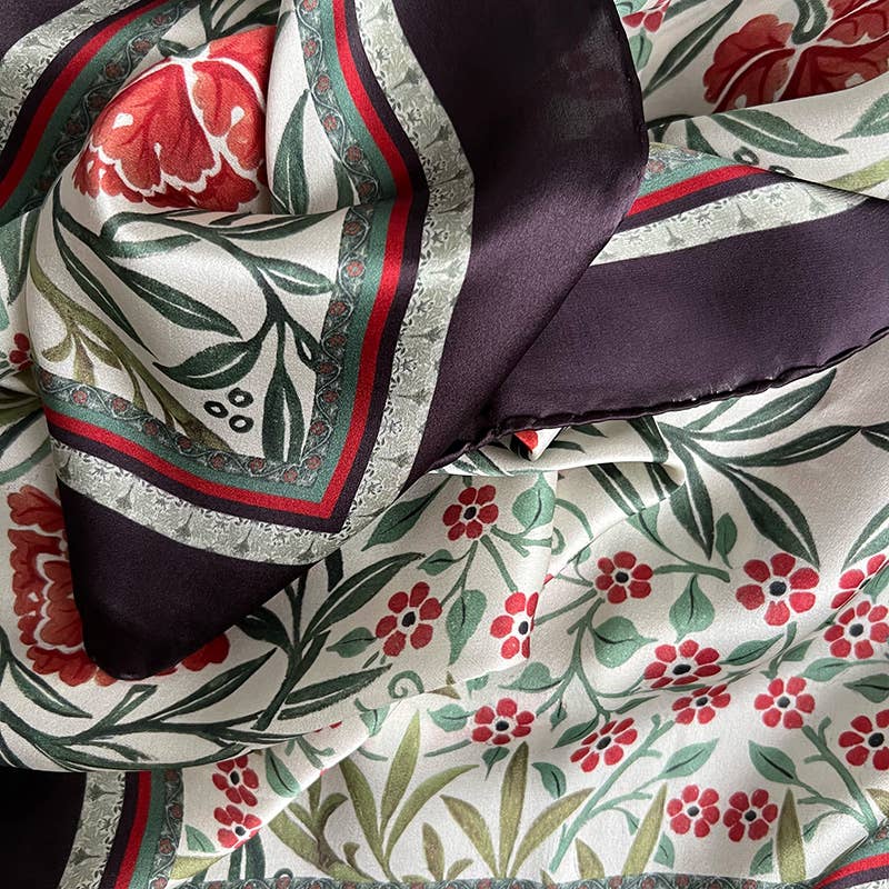 Eine Nahaufnahme des Koustrupco-Seidenschals „Morris Oxford“ Rot (90 x 90 cm) zeigt den seidigen Stoff mit roten Blumenmustern, grünen Blättern und geometrischen Bordüren in Oxford-Rot, der sanft und elegant im Damenstil gefaltet ist.