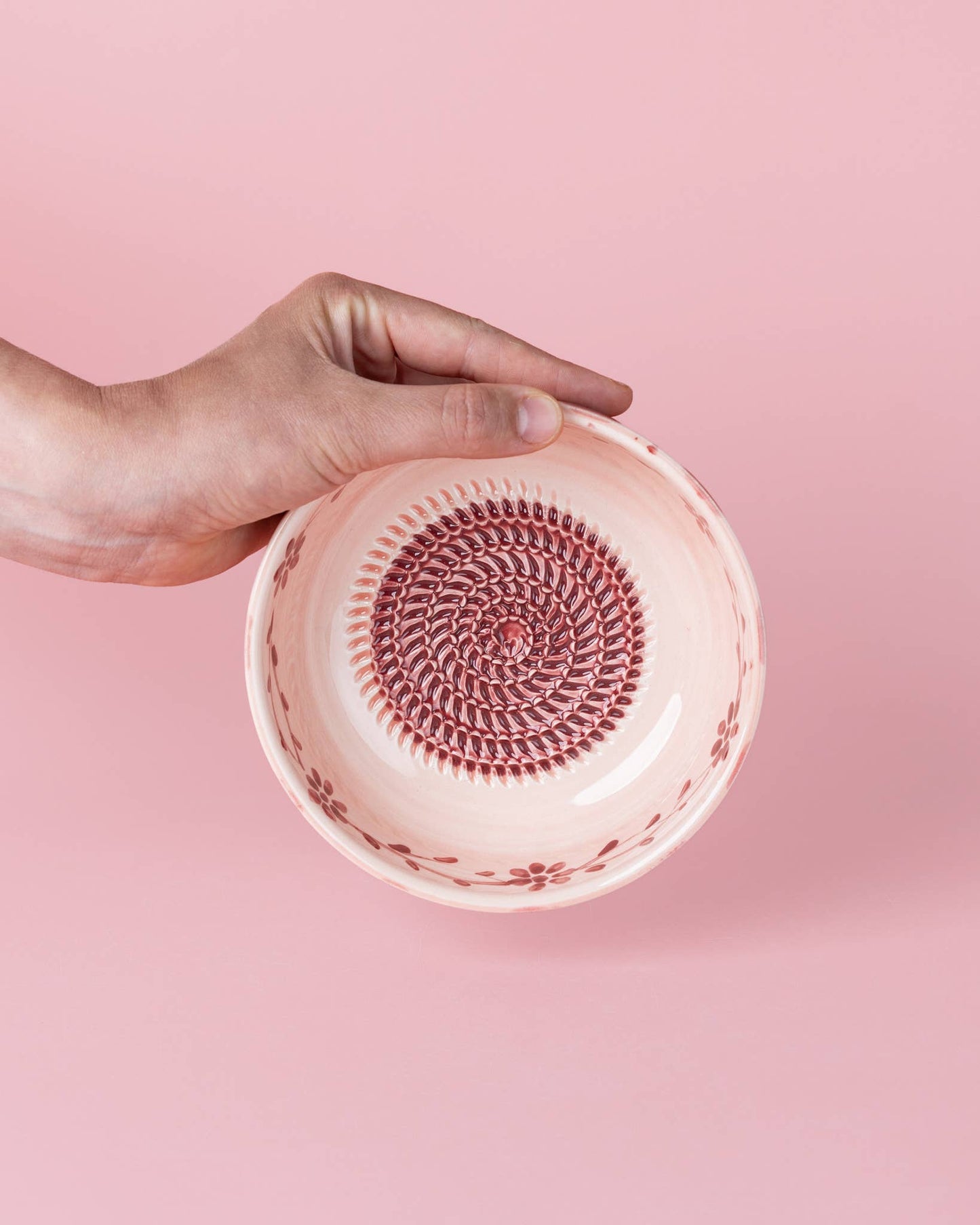 Eine Hand hält die Alfar Tierra Cocida Keramikschale mit Knoblauchreibe in Mittelrosa „Sakura“ – eine kleine Keramikschale, die von der traditionellen Keramik-Reibplatte inspiriert ist und sich durch ein zartes rosa Blumenmuster am Rand sowie eine kreisförmige, strukturierte Mitte auszeichnet, die die handbemalte spanische Handwerkskunst vor einem rosa Hintergrund zur Geltung bringt.