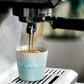 Eine Costa Nova 'Grespresso' Espressotasse Aqua aus Steingut und Keramik (9 cl) mit blau-weißem Wellenmuster steht auf einer Metallschale, in die eine Espressomaschine den Kaffee einfüllt.