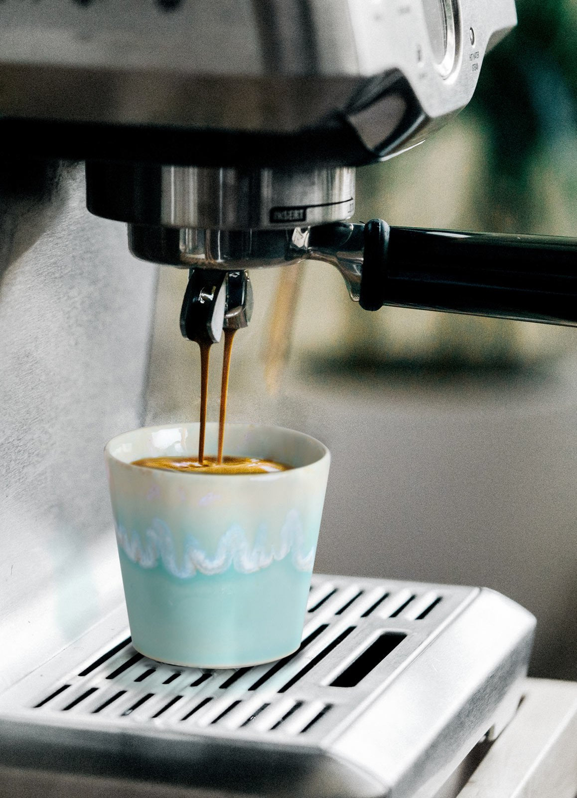 Eine Costa Nova 'Grespresso' Espressotasse Aqua aus Steingut und Keramik (9 cl) mit blau-weißem Wellenmuster steht auf einer Metallschale, in die eine Espressomaschine den Kaffee einfüllt.