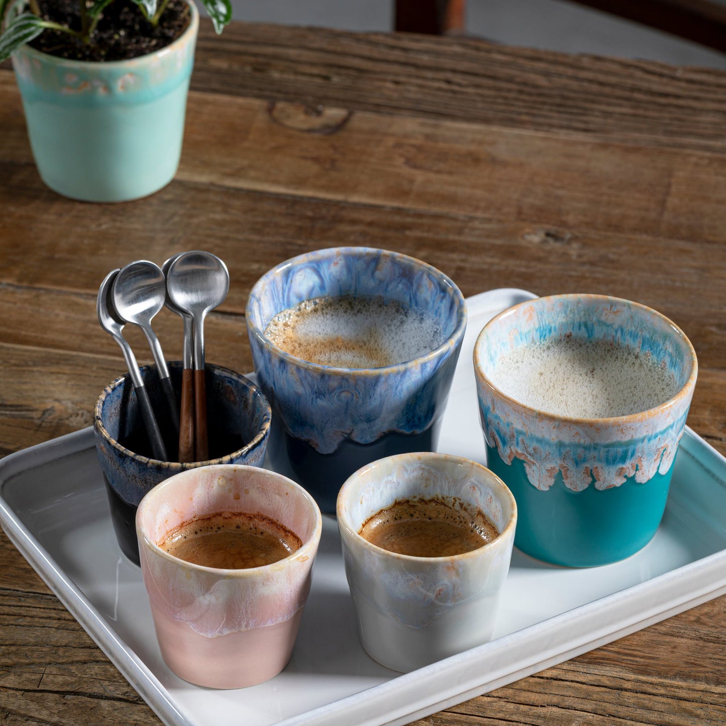 Ein Tablett mit fünf Keramikbechern, darunter eine Costa Nova 'Grespresso' Espressotasse Zartrosa aus Steingut und Keramik 9 cl, alle gefüllt mit Kaffee, sowie eine Tasse mit Metalllöffeln auf einem Holztisch mit einer Topfpflanze im Hintergrund.