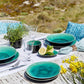 Auf einem Tisch im Freien stehen der 'Azurblau' Salatteller aus Keramik 21cm von Costa Nova, klare Gläser, Silberbesteck, ein Kräutertopf, Zitronen mit Basilikum, gelbe Blumen auf einem weißen Läufer auf einem rustikalen Holztisch. Gras und Pflanzen sorgen für eine frische Note.