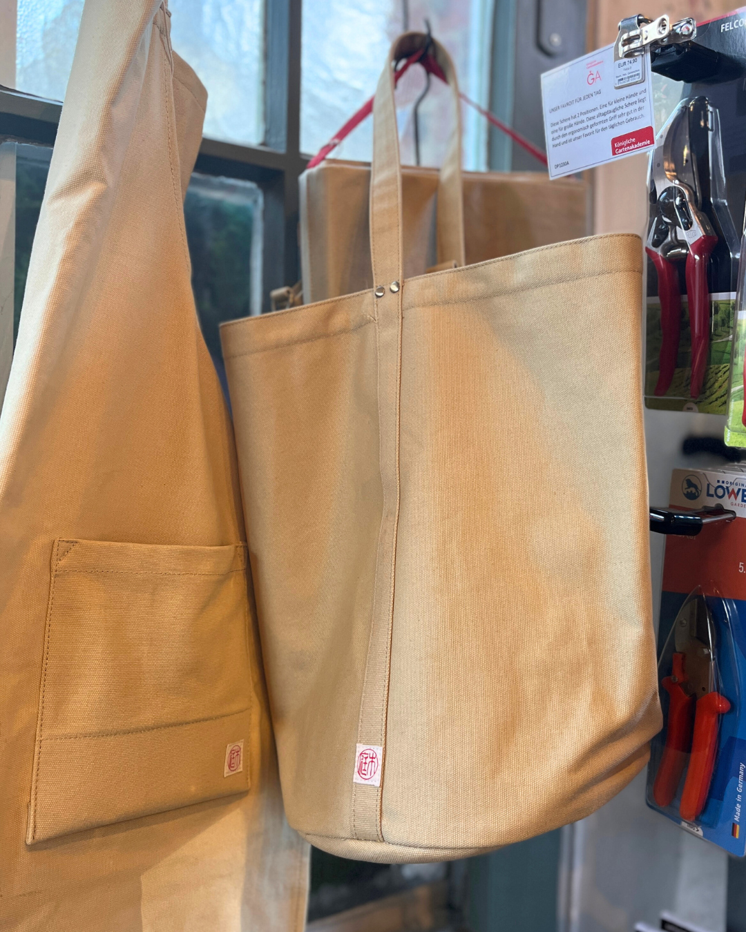 Eine hellbraune Kantan Werkzeugtasche - Niwaki und eine passende Schürze von Niwaki hängen in der Nähe eines Schaufensters. Die Tasche mit zwei Griffen und einem kleinen roten Logo ist robust und stilvoll. In der Nähe sind Gartengeräte ordentlich in einem Regal ausgestellt.