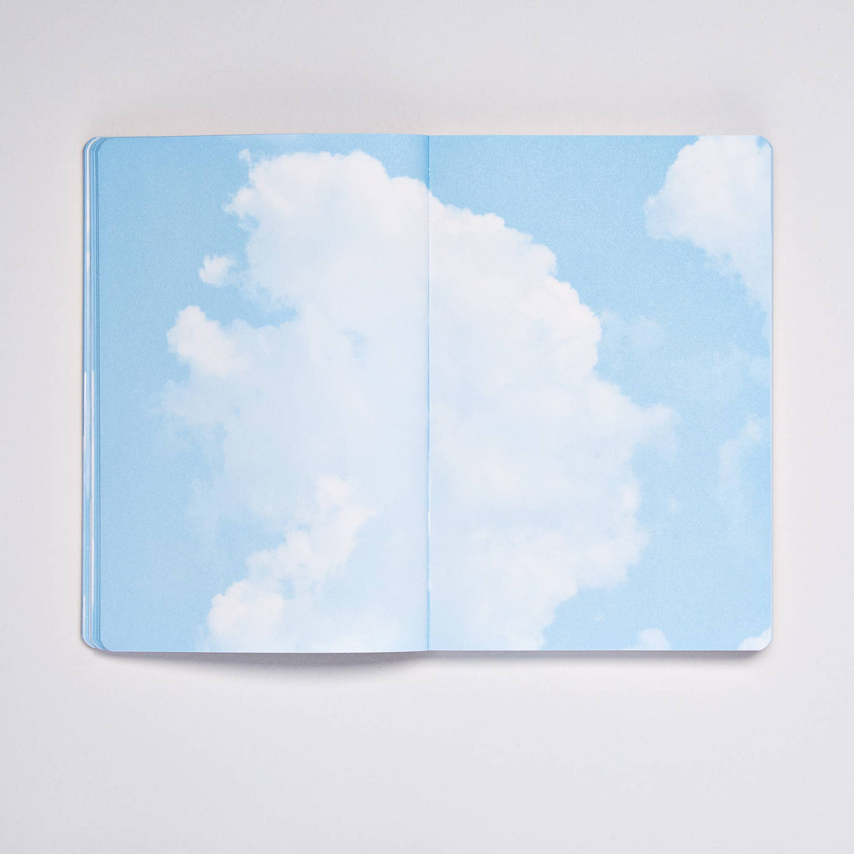 Ein aufgeschlagenes nuuna „Cloud Blue“-Inspirationsbuch in der Größe M von nuuna präsentiert 176 farbenfrohe Seiten, die mit einem blauen Himmel und flauschigen weißen Wolken bedruckt sind und so den Eindruck erwecken, man blicke in den Himmel – wodurch jeder Moment zu einem wahrhaft inspirierenden Erlebnis wird.