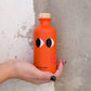 Eine Hand mit schwarz lackierten Nägeln hält eine orangefarbene Flasche Yiayia and friends | Natives Olivenöl extra mit Chili mit Cartoon-Augen und Holzkappe vor einer strukturierten weiß-grauen Putzwand.
