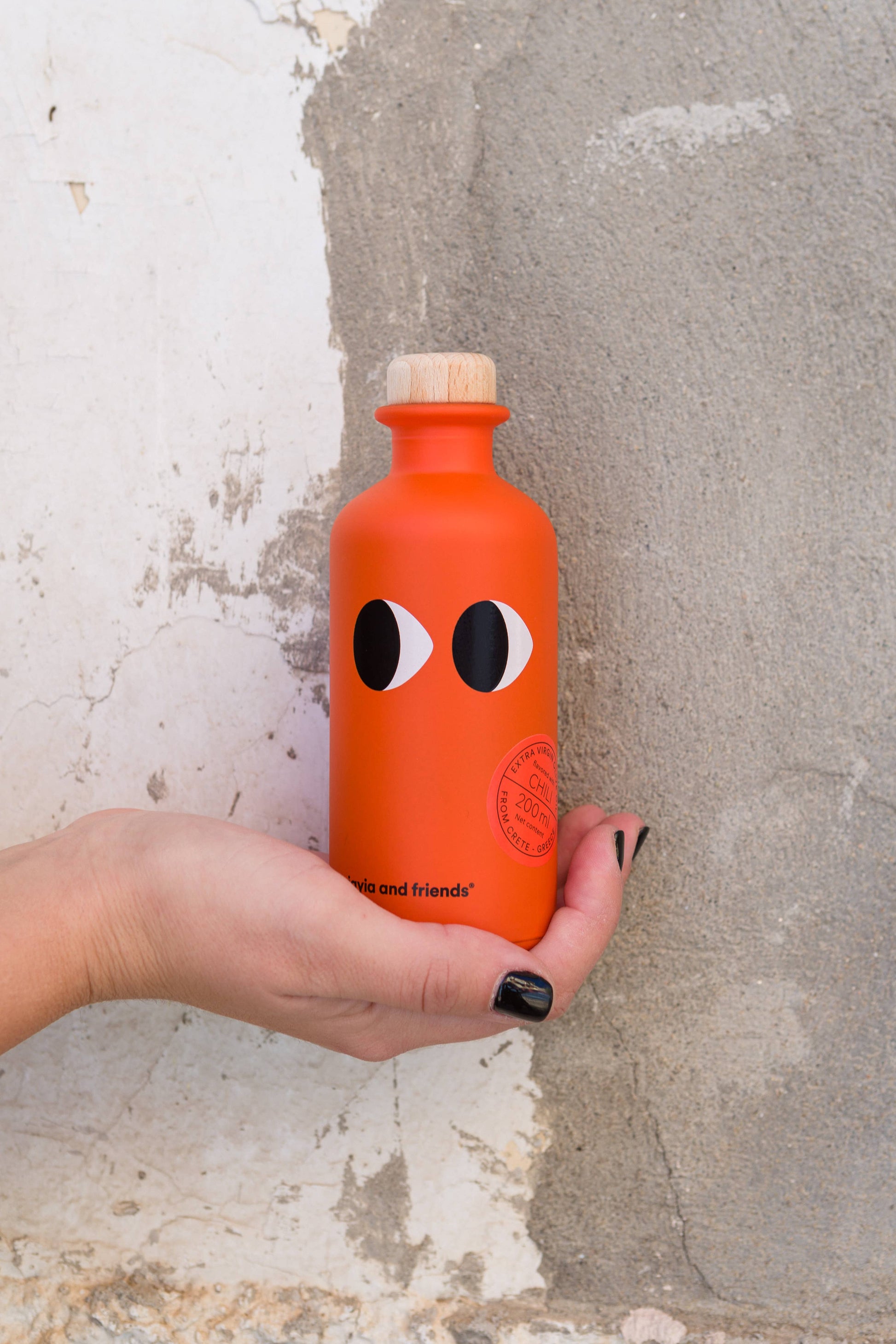 Eine Hand mit schwarz lackierten Nägeln hält eine orangefarbene Flasche Yiayia and friends | Natives Olivenöl extra mit Chili mit Cartoon-Augen und Holzkappe vor einer strukturierten weiß-grauen Putzwand.
