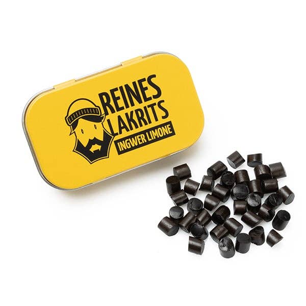 Eine gelbe LASSE LAKRITS-Dose mit der Aufschrift "Reines Lakritz Ingwer Limone" mit einem bärtigen Gesicht steht neben einem Stapel kleiner, schwarzer, zuckerfreier Lakritz auf weißem Grund.