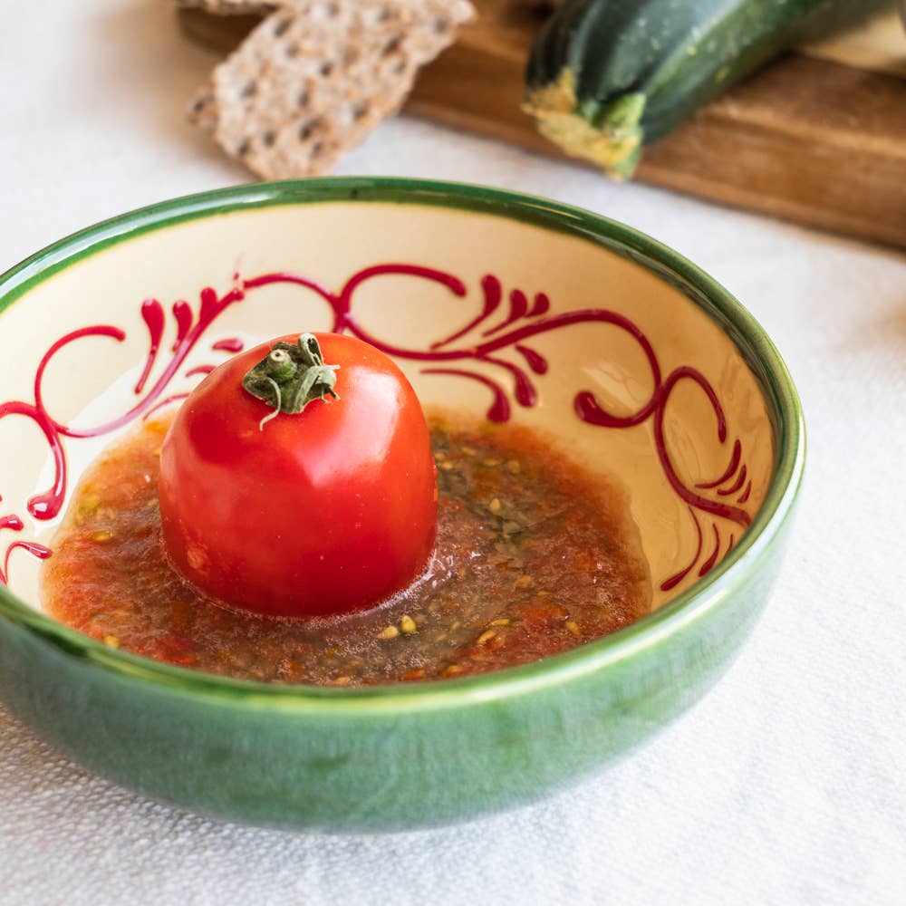 Eine Tomate sitzt in einer Suppe mit Kräutern, serviert in der grünen "Alhambra" Knoblauchreibe-Schale (15 x 15 x 5 cm) von Alfar Tierra Cocida; im Hintergrund sind Zucchini und Knäckebrot zu sehen.